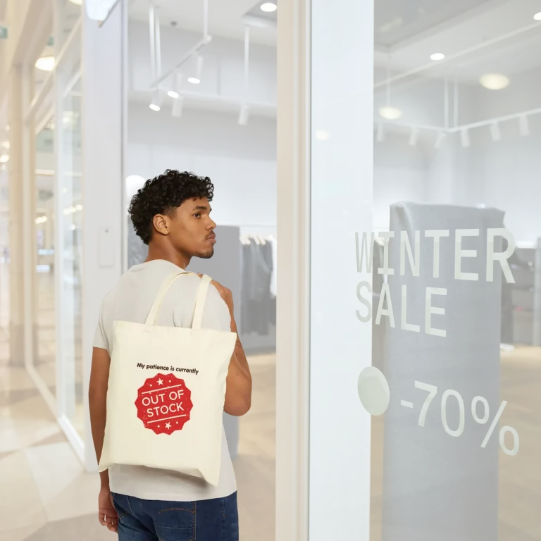 ⁠A man carrying a cream canvas tote bag over his shoulder that reads My Patience is Currently Out of Stock in a red stamp design.