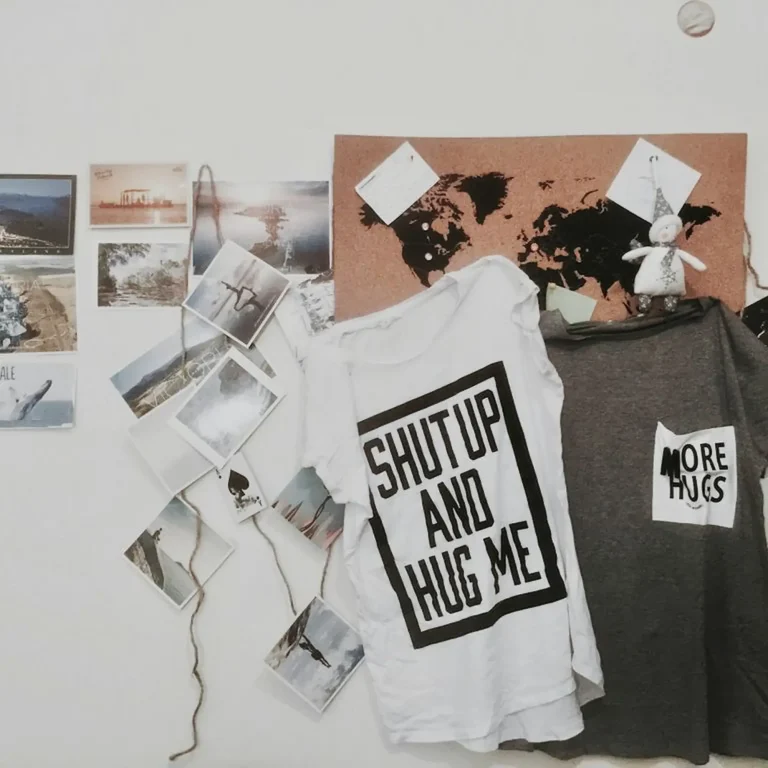 ⁠A white t-shirt reading Shut Up and Hug Me in a bold black box hanging next to a grey t-shirt with a white pocket patch reading More Hugs, 